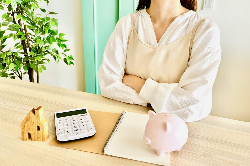 貯金箱と電卓、家計簿を前にして腕を組んで考える女性